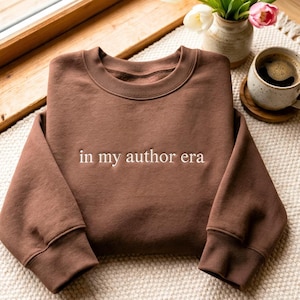 May include: A chocolate brown crewneck sweatshirt featuring the embroidered white text "in my author era" centred on the chest. The garment has long sleeves with ribbed cuffs and a ribbed neckline, constructed from a solid-coloured fabric.