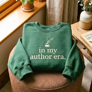 May include: A forest green crewneck sweatshirt featuring an embroidered white quill pen and inkwell graphic above the text "in my author era." The garment has long sleeves and a ribbed neckline, cuffs, and hem, displayed on a textured surface.