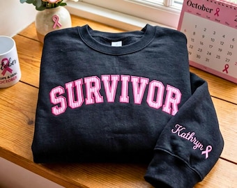 May include: A black crewneck sweatshirt featuring the word SURVIVOR embroidered in pink and white block lettering across the chest. The left sleeve includes the name Kathryn embroidered in white script above a small pink ribbon symbol.