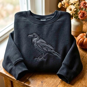 Könnte beinhalten: Ein schwarzes Sweatshirt mit Rundhalsausschnitt und einer detaillierten, einfarbigen Stickerei eines Raben im Profil. Der Vogel ist mittig auf der Brust platziert und zeigt eine komplexe Federstruktur sowie einen spitzen Schnabel in Dunkelgrautönen.
