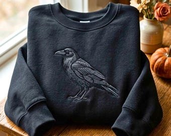 Peut inclure: Un sweat-shirt noir à col rond orné d'une broderie monochrome détaillée représentant un corbeau de profil. L'oiseau est placé au centre de la poitrine, mettant en valeur des textures de plumes complexes et un bec pointu, dans des tons gris foncé.