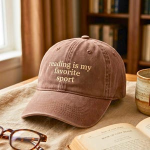 May include: A brown baseball cap with the text reading is my favorite sport embroidered in light beige thread on the front panel. The cap features a structured crown, a curved brim with stitched detailing, and eyelets for ventilation.