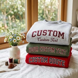 Puede incluir: Una pila de tres sudaderas de cuello redondo en gris claro, verde oliva y rojo. La sudadera gris presenta el texto CUSTOM Custom Text, la verde dice YOUR CUSTOM TEXT y la roja dice CUSTOM QUOTE en letras negritas de estilo universitario.