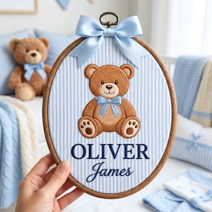 May include: An oval wooden embroidery hoop featuring a light blue and white striped fabric background. Centered is an embroidered brown teddy bear wearing a light blue bow. Below the bear, the names Oliver James are stitched in dark blue.