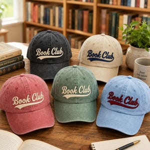 Puede incluir: Cinco gorras de béisbol en gris carbón, beige, rojo, verde salvia y azul claro, cada una con el texto Book Club bordado en letra cursiva en la parte frontal. Las gorras tienen un acabado desgastado y una corona estructurada con visera curva.