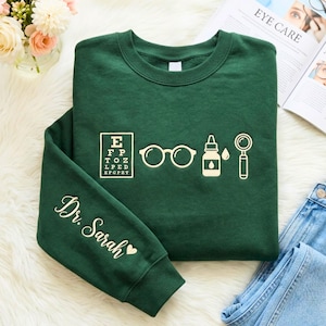 May include: A dark green crewneck sweatshirt featuring a cream-colored graphic of an eye chart, eyeglasses, an eye drop bottle, and a magnifying glass. The sleeve is printed with the text Dr. Sarah followed by a small heart symbol.