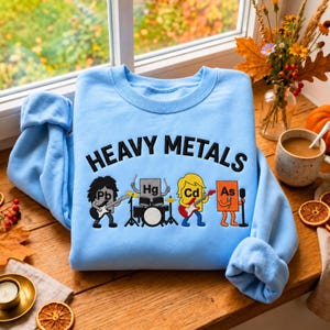 以下が含まれることがあります： 黒い文字で「HEAVY METALS」と書かれた水色のスウェットシャツ。テキストの下には、バンドとして描かれた4つの擬人化された化学元素記号があります。Pbはギター、Hgはドラム、Cdはギターを演奏し、Asはマイクに向かって歌っています。