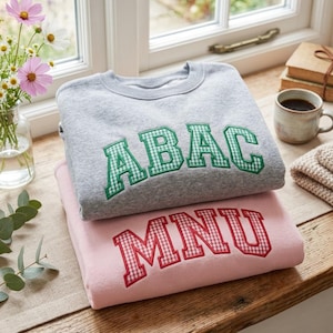 May include: A grey crewneck sweatshirt with green gingham applique letters spelling ABAC stacked on a light pink crewneck sweatshirt with red gingham applique letters spelling MNU. Both garments feature a classic collegiate font style.