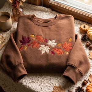 May include: A brown crewneck sweatshirt featuring an embroidered design across the chest. The embroidery depicts a cluster of autumn leaves in shades of deep burgundy, burnt orange, cream, and golden yellow, arranged in a horizontal pattern.