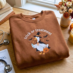 May include: A brown crewneck sweatshirt featuring an embroidered design of a white goose wearing a black witch hat and cape. The goose holds a blue syringe and an orange jack-o'-lantern bucket. The text "trick or treatment" is arched above.
