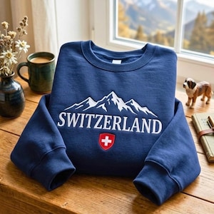 May include: A navy blue crewneck sweatshirt featuring a white embroidered graphic of mountain peaks above the text SWITZERLAND in white capital letters, with a small red shield emblem containing a white cross centered below the text.