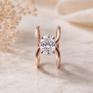 Peut inclure: Une bague en or rose avec une pierre précieuse transparente taille ovale au centre, maintenue par quatre griffes. L'anneau se compose de deux segments métalliques incurvés et parallèles qui s'arquent vers l'extérieur pour former un cadre ouvert.