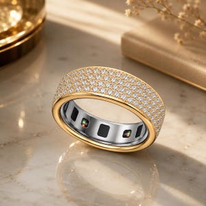 May include: A gold band ring featuring a wide surface paved with numerous small, clear, round-cut stones. The interior of the ring is silver-toned with a series of black rectangular insets and small electronic sensor components visible inside.