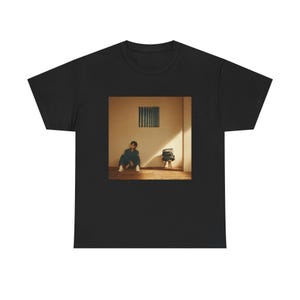 May include: A black t-shirt featuring a centred rectangular graphic print. The image depicts a person sitting on a wooden floor in a room with beige walls, a small stool with stacked items, and a vertical striped wall art piece above them.