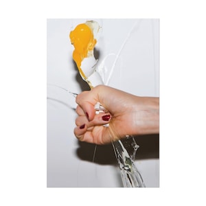 May include: A fist tightly gripping a raw egg, causing the white shell to crack and shatter. The bright yellow yolk and clear egg white are spilling out and dripping downward, creating a dynamic motion effect against a plain white background.