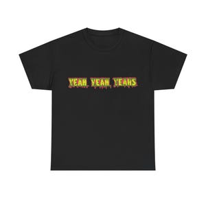 Op de afbeelding: Een zwart T-shirt met korte mouwen en de tekst YEAH YEAH YEAHS op de borst gedrukt. De letters zijn uitgevoerd in een heldere gele kleur met een textuur en druipend effect, omlijnd met een donkerrode rand op de zwarte stof van het shirt.