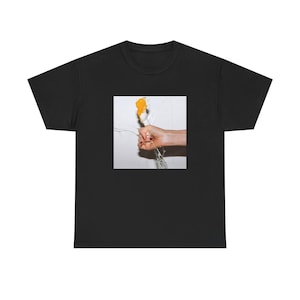 Op de afbeelding: Een zwart t-shirt met een vierkante grafische print op de borst. De afbeelding toont een hand die een doorzichtige glazen fles vastgrijpt die kapot gaat, waarbij een feloranje vloeistof tegen een effen witte achtergrond spat.