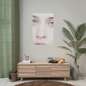 a wooden cabinet with a mirror above it, a small deer figurine, and a plant in a vase. The cabinet has a mirror on the front and a small deer figurine on top. The plant in the vase is green and has long leaves. The wall behind the cabinet has a poster of a woman's face.