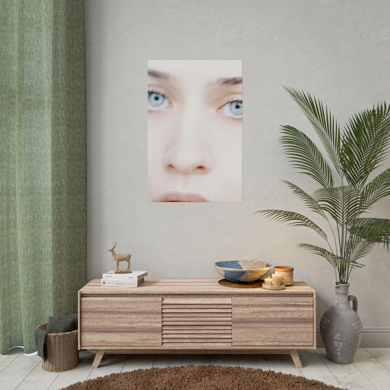 a wooden cabinet with a mirror above it, a small deer figurine, and a plant in a vase. The cabinet has a mirror on the front and a small deer figurine on top. The plant in the vase is green and has long leaves. The wall behind the cabinet has a poster of a woman's face.
