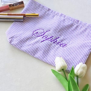 May include: A purple and white seersucker zippered pouch featuring the name Sophia embroidered in purple script lettering. The pouch is accompanied by two white artificial tulips and three cosmetic items including a lip gloss and pencils.
