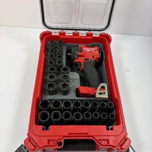 Può includere: Una valigetta rossa Milwaukee Packout con coperchio trasparente contenente un avvitatore a impulsi a batteria rosso e nero e una serie di bussole a impatto in metallo nero disposte in un inserto grigio sagomato all'interno del vano.