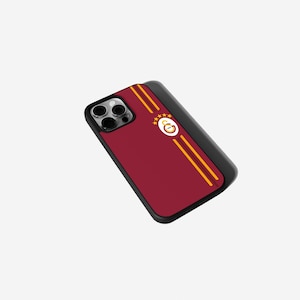 Op de afbeelding: Een bordeauxrode telefoonhoes met een zwarte rand. De hoes heeft twee verticale gouden strepen en een cirkelvormig embleem met een sterrenontwerp. De telefoonhoes is ontworpen voor een smartphone.