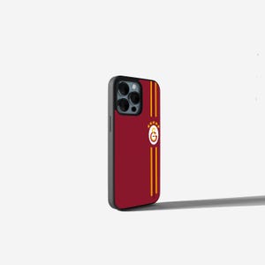 Op de afbeelding: Een bordeauxrode telefoonhoes met een zwarte rand. De hoes heeft twee verticale gouden strepen en een cirkelvormig embleem met een gouden omtrek en vier gouden sterren. De hoes is ontworpen voor een smartphone.