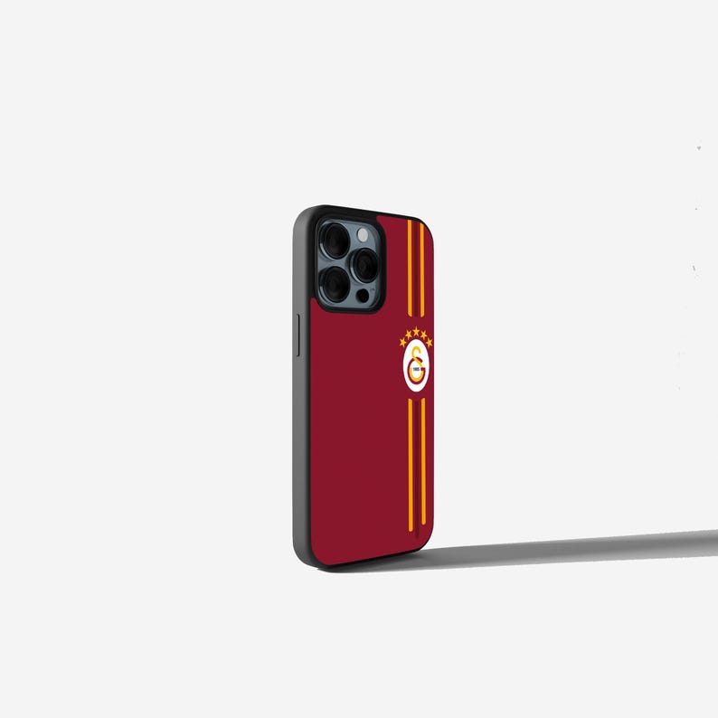 Op de afbeelding: Een bordeauxrode telefoonhoes met een zwarte rand. De hoes heeft twee verticale gouden strepen en een cirkelvormig embleem met een gouden omtrek en vier gouden sterren. De hoes is ontworpen voor een smartphone.