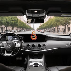 May include: Interior view of a Mercedes-Benz car with a Galatasaray football club logo hanging from the rearview mirror. The logo is orange, red, and yellow. The car's dashboard is black with silver accents, and the steering wheel is on the left.