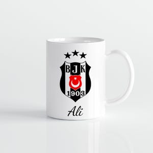 May include: White ceramic mug featuring the Besiktas JK emblem, a black and white shield with a red crescent moon and star. The mug also has the text "Ali" in black script.