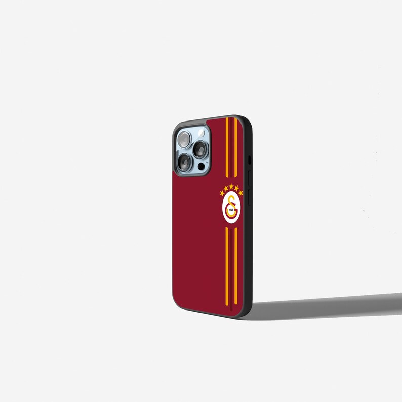 Op de afbeelding: Een bordeauxrode telefoonhoes met een zwarte rand. De hoes heeft twee verticale gouden strepen en een logo met een rood-gouden embleem en drie gouden sterren. De hoes is ontworpen voor een smartphone.
