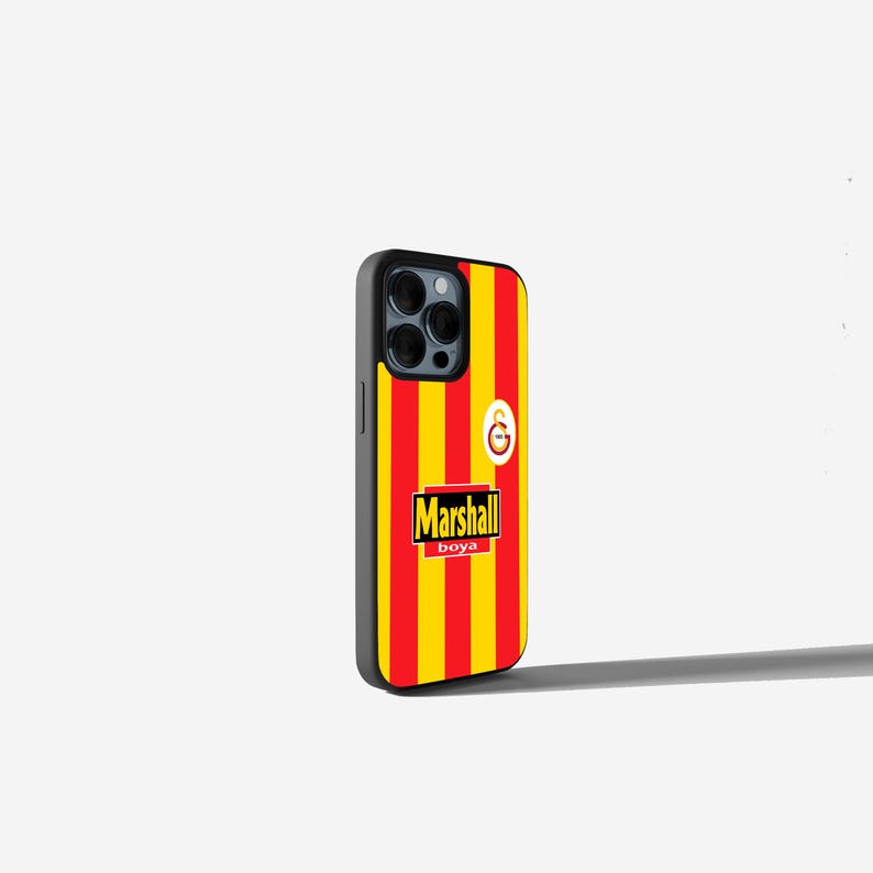 May include: A black phone case with a red and yellow striped design, featuring the text "Marshall boya" and a team logo. The case has a black border and covers the phone's camera.