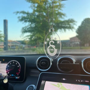 Könnte beinhalten: Transparenter, ovaler Autoanhänger mit einer silbernen Kette. Der Anhänger zeigt die Nummer "1905" und ein Logo mit Sternen und dem Buchstaben "G" in einer Halbmondform. Der Anhänger hängt vor dem Armaturenbrett eines Autos.