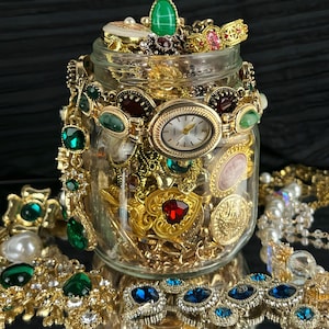 May include: A clear glass jar filled with an assortment of gold-toned jewelry, including necklaces, bracelets, and a wristwatch with a white face. The collection features various colorful gemstones, such as green, red, and blue stones, along with pearls.