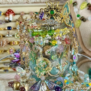 Könnte beinhalten: Ein klares Glasgefäß gefüllt mit einer Auswahl an buntem Schmuck, darunter goldfarbene Ketten, Perlenketten und schmetterlingsförmige Anhänger. Das Gefäß ist mit einem goldenen Schmetterlingsornament mit einem grünen Mittelstein verziert.
