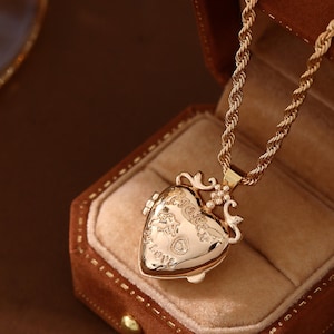 Könnte beinhalten: Eine goldfarbene herzförmige Medaillon-Halskette mit einer gedrehten Seilkette. Das Medaillon ist mit dem Wort "Love" und anderen dekorativen Elementen graviert. Die Halskette wird in einer braunen Samt-Schmuckschatulle präsentiert.