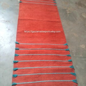 May include: A rectangular woven runner rug in a solid burnt orange color featuring horizontal stripes of light blue thread. The rug has light blue fringe tassels along the edges. The text https://gautamartexports.etsy.com is printed in the center.
