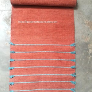 May include: A rectangular terracotta-colored woven runner rug featuring horizontal light blue stripes and matching blue tassels along the sides. The text https://gautamartexports.etsy.com is printed in white on the upper section of the rug.