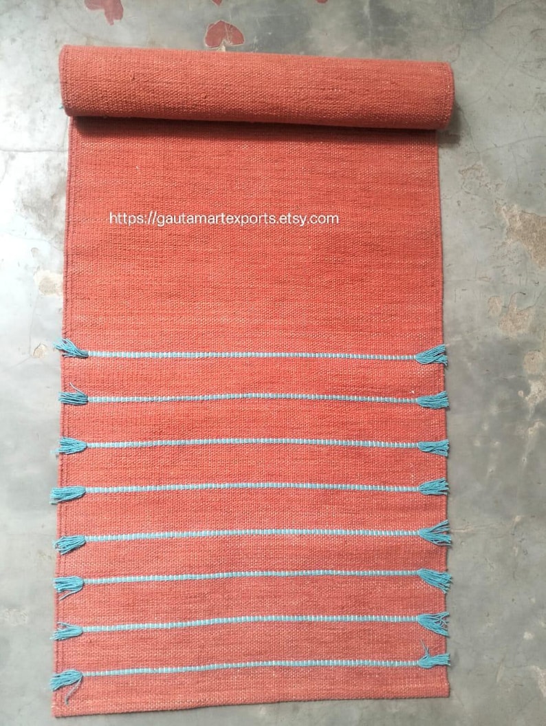 May include: A rectangular terracotta-colored woven runner rug featuring horizontal light blue stripes and matching blue tassels along the sides. The text https://gautamartexports.etsy.com is printed in white on the upper section of the rug.