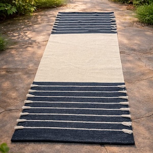 May include: A rectangular rug with a cream-coloured centre and navy blue ends. The navy sections feature horizontal white stripes and tassels. The rug is placed on a stone surface, with greenery in the background.