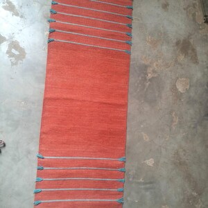 May include: A rectangular orange table runner featuring horizontal light blue stripes at both ends. The runner has a woven texture and includes small, light blue fringe tassels attached to the edges of each horizontal stripe along the sides.