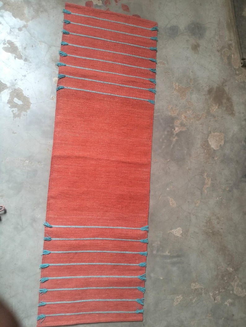 May include: A rectangular orange table runner featuring horizontal light blue stripes at both ends. The runner has a woven texture and includes small, light blue fringe tassels attached to the edges of each horizontal stripe along the sides.