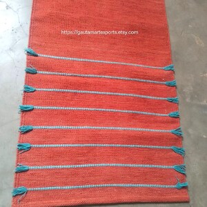 May include: A rectangular orange table runner featuring a woven texture and eight horizontal stripes in light blue. Each light blue stripe ends with small tassels on both sides. The text https://gautamartexports.etsy.com is printed in white.