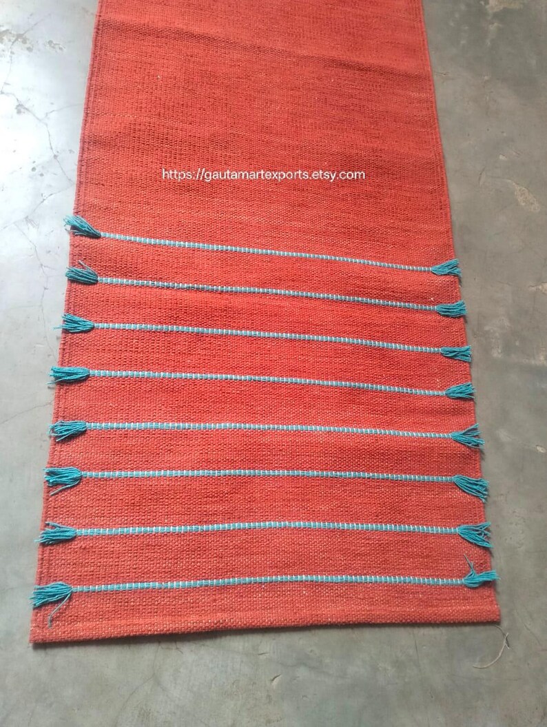 May include: A rectangular orange table runner featuring a woven texture and eight horizontal stripes in light blue. Each light blue stripe ends with small tassels on both sides. The text https://gautamartexports.etsy.com is printed in white.
