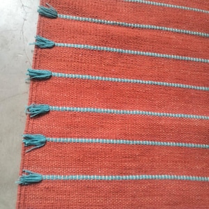 May include: A woven rug in a solid burnt orange color featuring horizontal stripes in a light blue thread. The left edge of the rug is finished with a series of light blue tassels attached to the end of each horizontal stripe.
