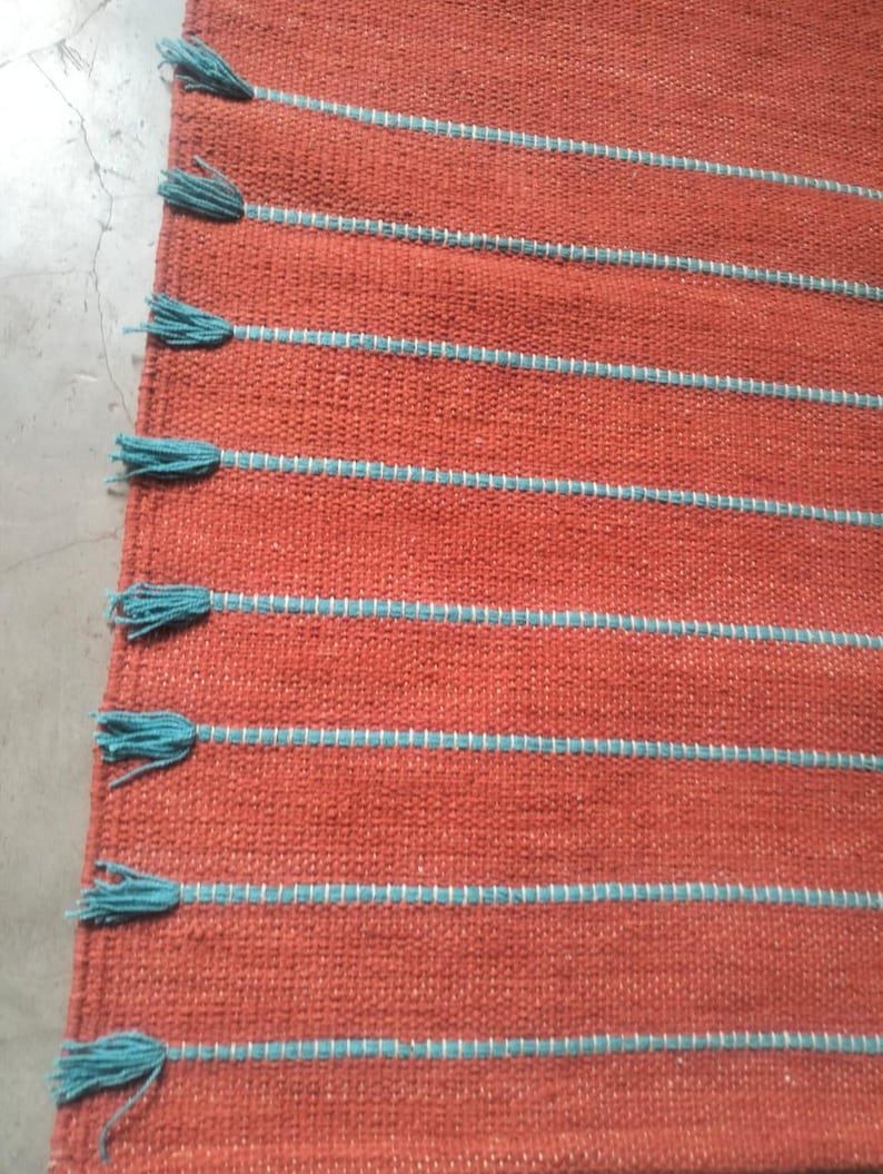 May include: A woven rug in a solid burnt orange color featuring horizontal stripes in a light blue thread. The left edge of the rug is finished with a series of light blue tassels attached to the end of each horizontal stripe.