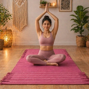 May include: A person in a pink yoga outfit sits cross-legged on a magenta yoga mat with a fringed edge, arms raised in prayer. The room has wooden floors, plants, and decorative baskets, creating a serene atmosphere for yoga and meditation.