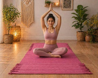 May include: A person in a pink yoga outfit sits cross-legged on a magenta yoga mat with a fringed edge, arms raised in prayer. The room has wooden floors, plants, and decorative baskets, creating a serene atmosphere for yoga and meditation.