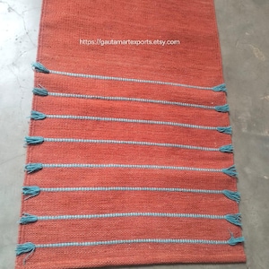 May include: A rectangular orange table runner featuring a woven texture and eight horizontal light blue stripes. Each stripe ends with small light blue tassels on both sides. The text https://gautamartexports.etsy.com is printed in white.