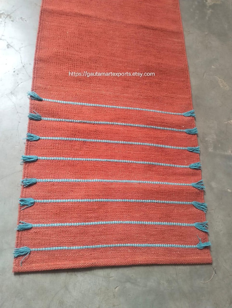 May include: A rectangular orange table runner featuring a woven texture and eight horizontal light blue stripes. Each stripe ends with small light blue tassels on both sides. The text https://gautamartexports.etsy.com is printed in white.